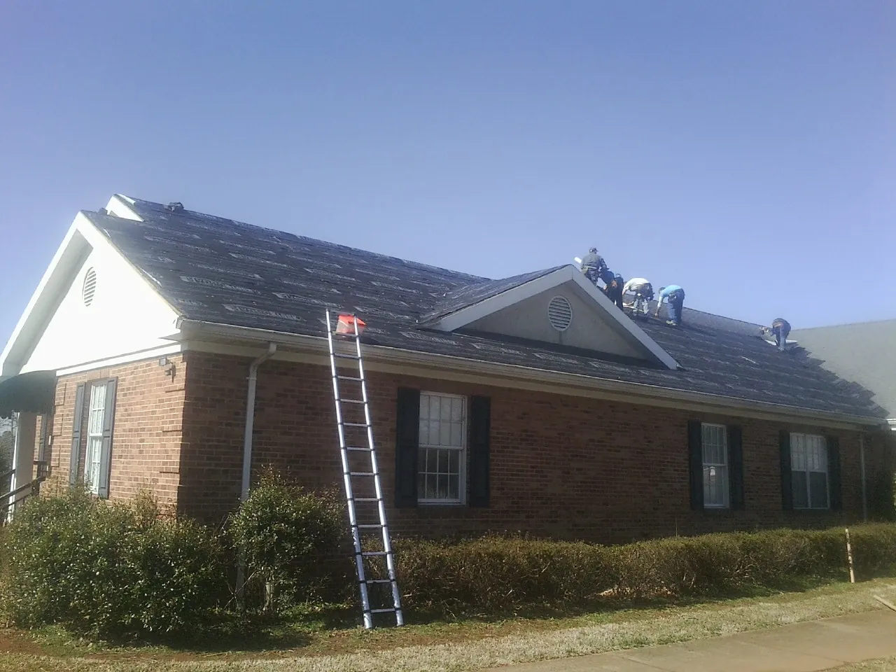 Crew tearing shingles off a brick ranch roof with a ladder set — Metal Roof Repair work by our crew in Intercession City